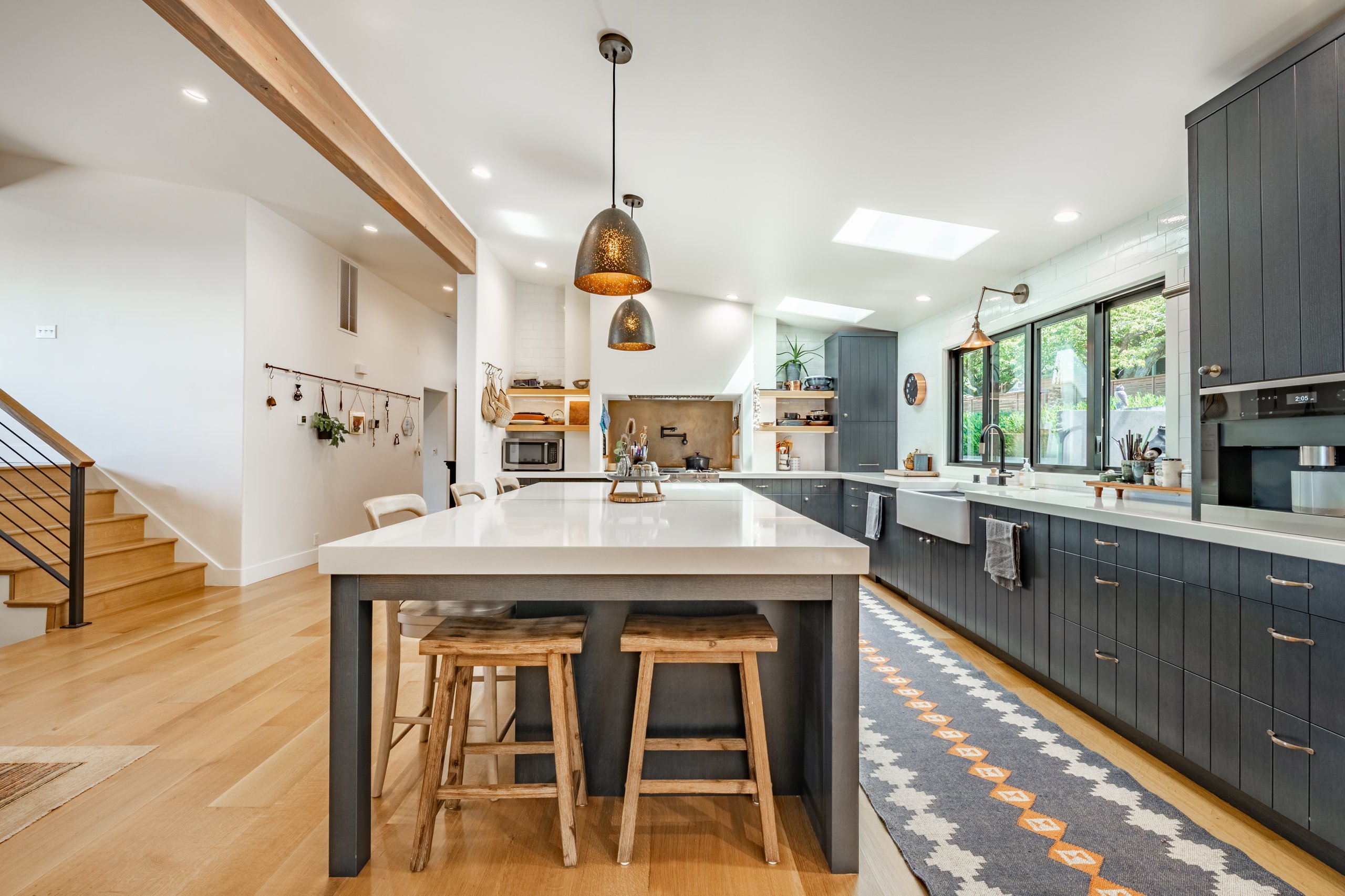 A modern kitchen with dark cabinetry, light countertops, warm wood flooring, a large central island with white countertop and wooden stools, and pendant lighting.