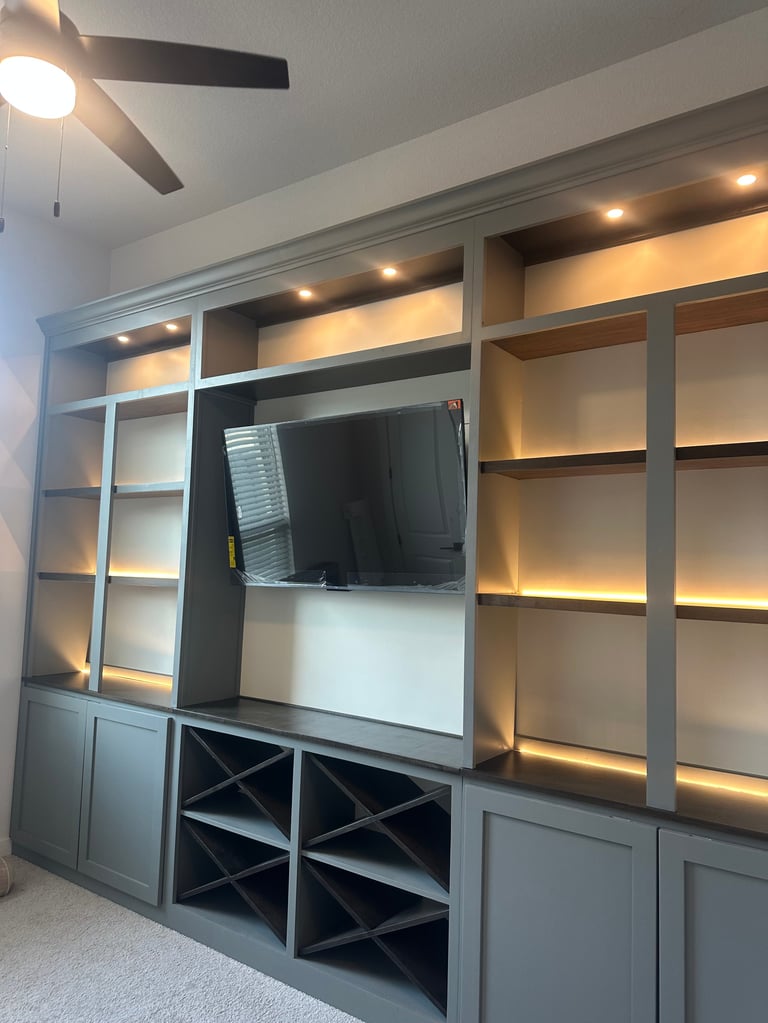 Modern gray built-in entertainment center with wall-mounted TV, illuminated shelving, wine racks, and ceiling fan in a contemporary bedroom