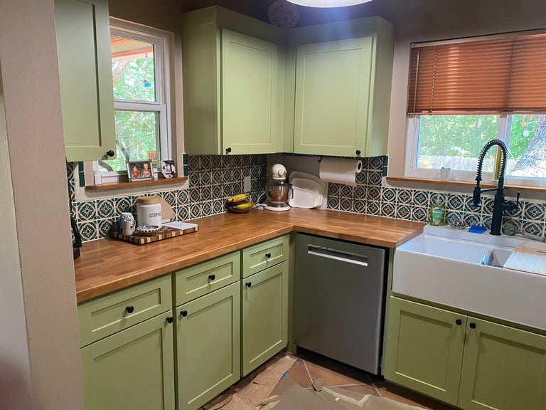 Green kitchen with wooden countertops, geometric tile backsplash, stainless steel appliances, and windows overlooking greenery