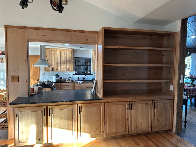 Modern kitchen with wooden cabinetry, open shelving, and a view into the main kitchen area through a large window opening